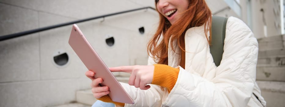 Young stylish girl, redhead female students sits on stairs outdoors with digital tablet, reads, uses social media app on gadget, plays games while waits on street.