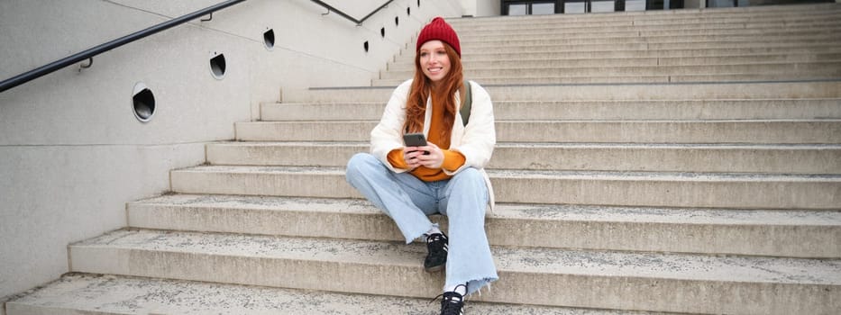 City lifestyle and mobile phone. Smiling college girl with red hair, sits on public stairs on street with smartphone, reads message, uses social media app.