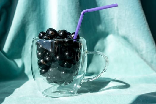 Summer fresh black currant berries in a glass mug with a cocktail tube on a turquoise fabric background. Sweet ripe black currant. Delicious healthy food, rich in vitamins. Organic Natural healthy food