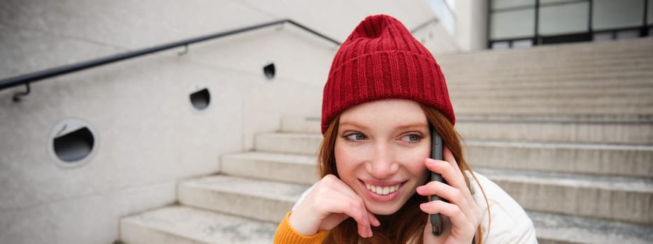 Beautiful smiling redhead female model, sits on street and talks on mobile phone, uses smartphone app to call abroad, laughing during telephone conversation.