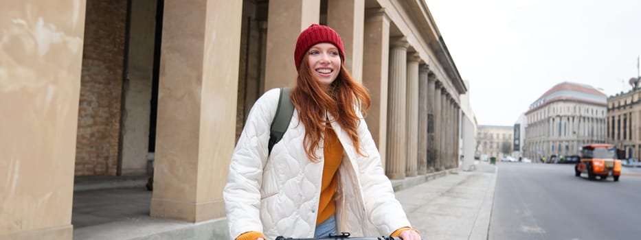 Smiling redhead european girl drives public e-scooter, tourist explores city, rides in city centre.