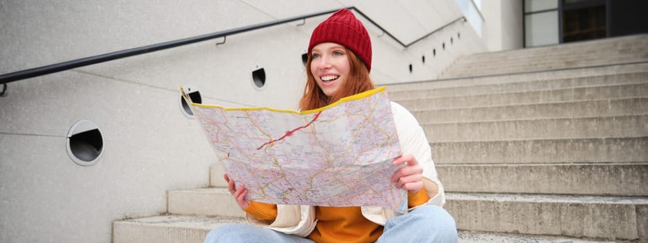 Young smiling redhead girl, tourist sits on stairs outdoors with city paper map, looking for direction, traveller backpacker explores city and looks for sightseeing.