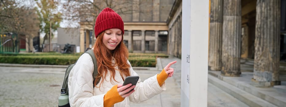 Tourism and sightseeing concept. Young redhead woman, tourist walks around city, looks at her smartphone app and at history stand, explores adventures.