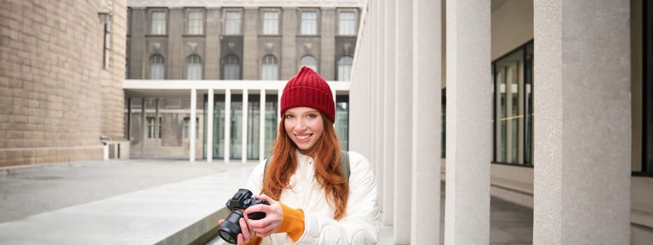 Smiling redhead girl photographer, taking pictures in city, makes photos outdoors on professional camera. Young talent and hobby concept