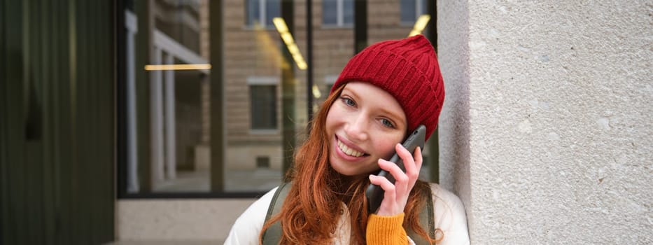 Happy beautiful girl, redhead tourist talks on mobile phone, making a call abroad, has conversation on smartphone, standing with backpack outdoors.