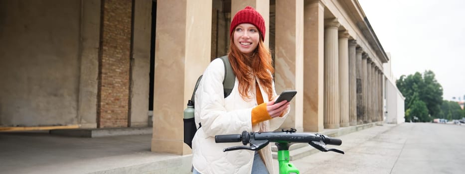 Beautiful young woman, tourist with backpack, rents e-scooter to travel around town, uses mobile phone application.