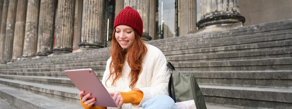 Outdoor shot of young stylish redhead girl sits on staircase and connects to public wifi, uses digital tablet, reads news on gadget.