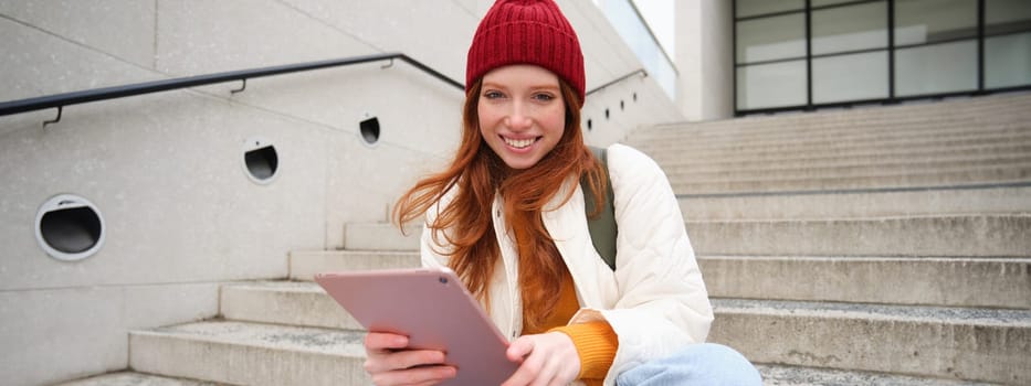 Young beautiful girl with long red hair, smiles, uses internet application, connect to wifi near campus, sits on stairs with digital tablet.