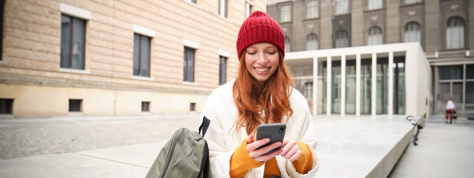 Young redhead woman with smartphone, sitting outdoors with backpack, student looking at her mobile phone, texts message.