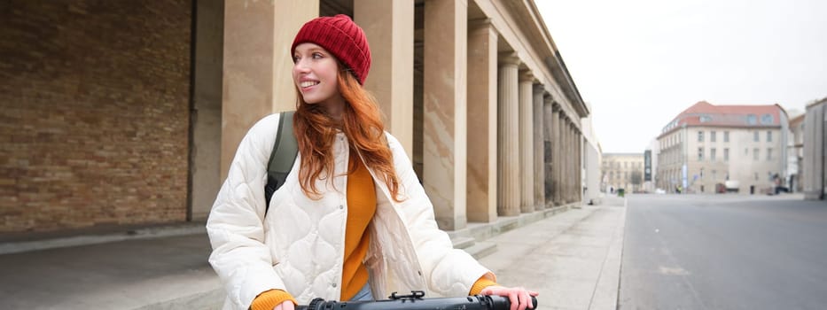 Young smiling redhead girl, student rides electric scooter, rents it and travels around city. Transport and travelling concept