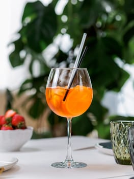 Summer drink Aperol Spritz aperitif served in wine glass with aperol, prosecco, soda, slice of orange. Refreshing drink Aperol Spritz cocktail on table in interior with green monstera plant background