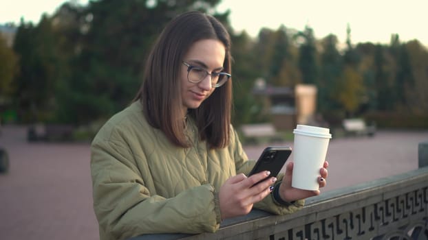 A young woman is chatting on a smartphone with a glass of coffee in her hand. A girl in glasses with a phone near the fence on the embankment with a hot drink in a glass. 4k