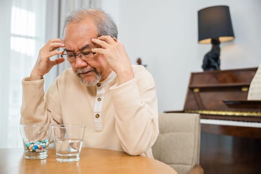 Asian senior old man grey haired sad sitting in chair hand hold head with many medicines and pills for taking, elderly man sick have headache looking pill glass on at home, Elderly healthcare