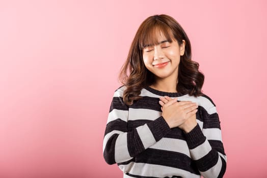Happy woman in casual clothes holding hands at chest close to heart meditating and smiling in studio short isolated on pink background, believe faith and love gratitude concept