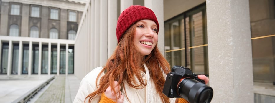 Smiling redhead girl photographer, taking pictures in city, makes photos outdoors on professional camera. Young talent and hobby concept