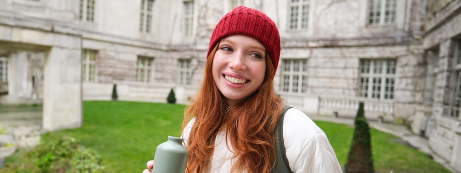 Portrait of happy young woman, tourist with backpack sightseeing, drinking hot tea from thermos, holding flask and smiling. Tourism and people concept