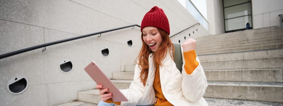 Happy stylish redhead girl, student in red hat, holds digital tablet, uses social media app, searches something online, connects to wifi.