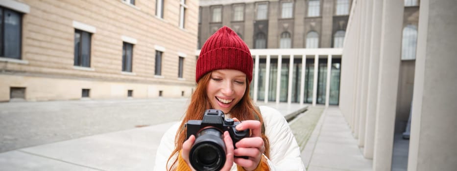 Smiling redhead girl photographer, taking pictures in city, makes photos outdoors on professional camera. Young talent and hobby concept