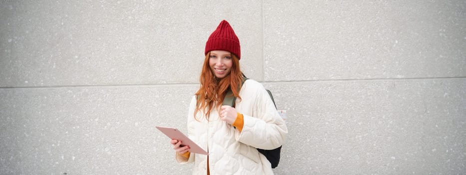 Young redhead woman with red hat, uses her digital tablet outdoors, stands on street with gadget, connects to wifi internet and searches for a location in internet.