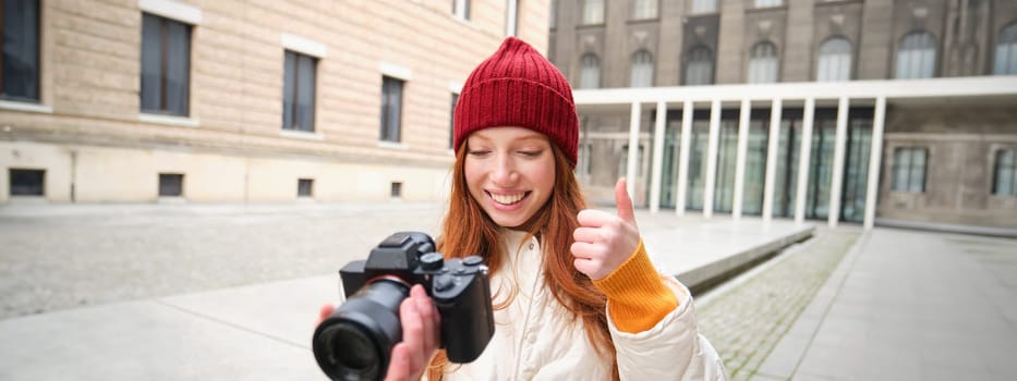 Smiling redhead girl photographer, taking pictures in city, makes photos outdoors on professional camera. Young talent and hobby concept