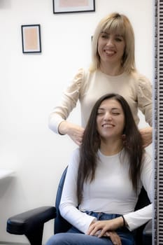 Beautiful young caucasian girl blonde hairdresser with a smile shows the end result pulling her hair to the face of a satisfied client sitting in a chair reflecting in the mirror,close-up side view and selective focus.Hairshop concept.