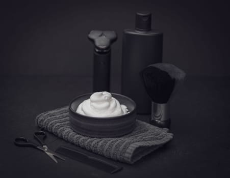 Shaving foam in a bowl with a terry towel, a comb, scissors, a shaving brush, an electric razor and a bottle of aftershave cream on a black background, close-up side view. The concept of a male hairdresser, a beauty salon, a beard shave.