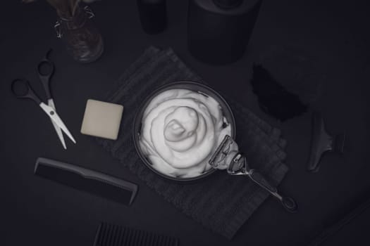 Shaving foam in a bowl with a terry towel, razor, comb, scissors, shaving brush, bar soap, electric razor, bottle of aftershave cream on a black background, flat lay close-up. The concept of a male hairdresser, beauty salon, beard shaving.