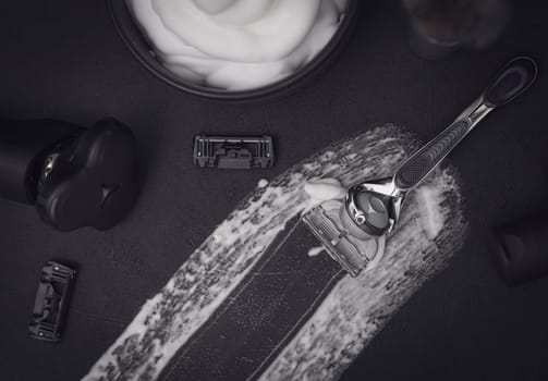 A smear of shaving foam in the center on a black background with a razor, a comb, a bottle of aftershave cream, a bowl of foam and an electric razor with copy space, flat lay close-up. The concept of a male hairdresser, beauty salon, beard shaving.