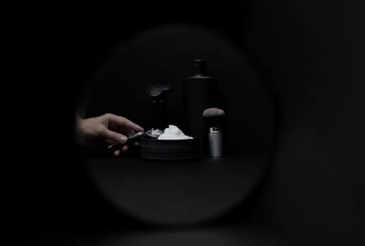 Shaving foam in a bowl, a shaving brush, an electric razor, a bottle of aftershave cream and a female hand holding a razor in the reflection of a round mirror on a black background, close-up side view. The concept of a male hairdresser, beauty salon, beard shaving.