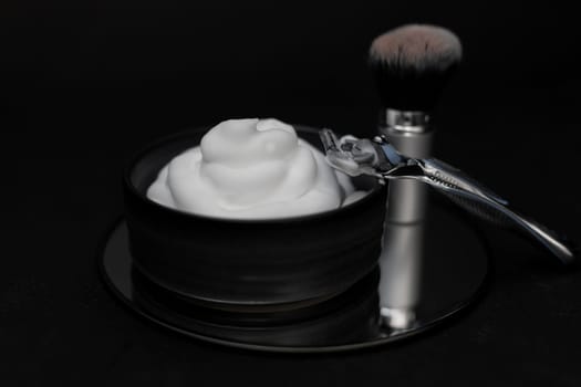 Shaving foam in a bowl, a shaving brush and a razor on a round mirror on a black background, close-up side view. The concept of a male hairdresser, beauty salon, beard shaving.