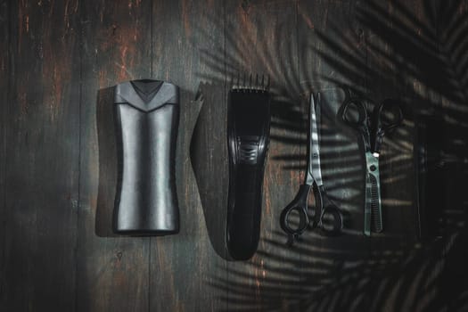 Scissors, combs, shampoo and men's hair clipper lie on a black homemade wooden background with a shadow from a palm branch, flat lay close-up. The concept of a male barbershop.