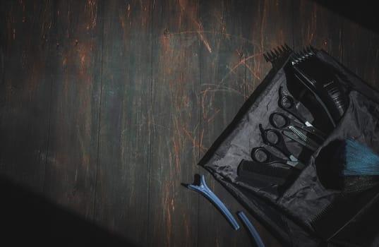 Scissors, combs, hair clipper, brush and clips with apron lie on the right on a black homemade wooden background with a little copy space on the left, flat lay close-up. The concept of a male barbershop, dark style.