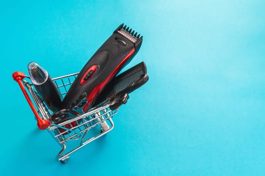 Men's haircut machine, scissors and comb lie in a mini cart on the left on a blue background with copy space on the right, close-up side view. The concept of hairdressing and shopping.