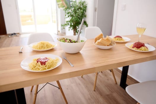 White plates with Italian spaghetti with fresh tomato sauce and a bowl of fresh healthy vegan salad, served on a wooden table in modern interior of a home living room. Traditional Italian cuisine.