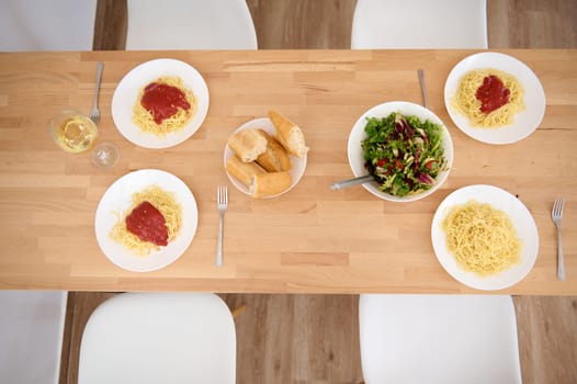 Top view of plates with appetizing spaghetti and classic tomato sauce, a wine glass of white wine and a bowl of fresh raw vegan salad on wooden table . Still life with traditional Italian food - pasta