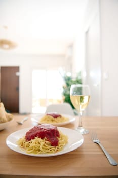 A plate with appetizing spaghetti and classic tomato sauce and a wine glass of white wine on the table at home interior. Still life with traditional Italian food - pasta. Copy advertising space