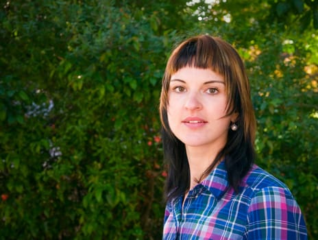 Side head and shoulders portrait of a young brunette woman in front of a green hedge in summer and looking at camera