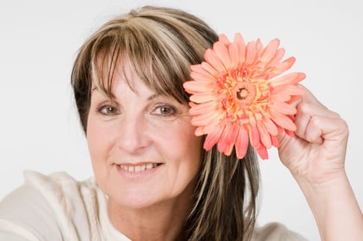 Head and shoulders portrait of middle-aged woman posing with orange-colored flower against neutral bright studio background
