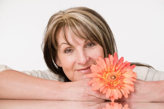 Head and shoulders portrait of a middle-aged woman with arms resting broadly on a table and orange flower against neutral light studio background