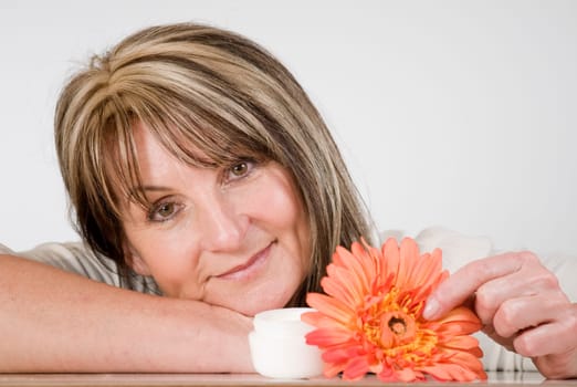 Head and shoulders shot of middle-aged woman with head resting on hands and cream jar and orange-colored artificial flower