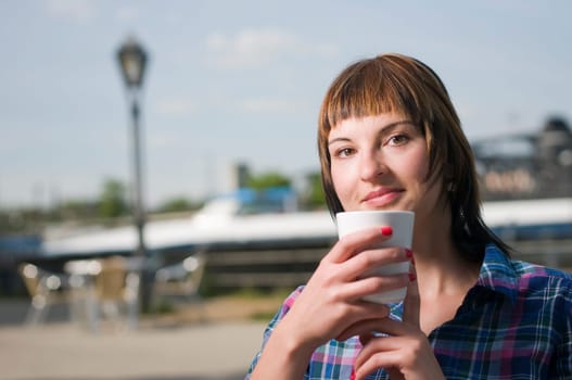 Head and shoulders shot of a young brown-haired woman sitting at a table in a street café and holding a white cup