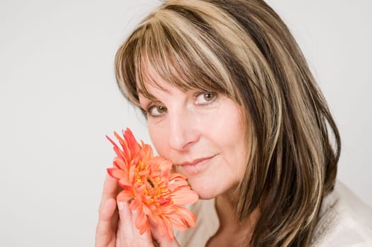 Head and shoulders portrait of middle-aged woman posing with orange-colored flower against neutral bright studio background