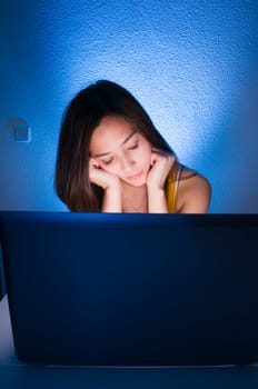 Head-and-shoulders portrait of a young black-haired woman sitting in front of the screen with her head resting on her hands,sleeping in front of blue backbone  Monitor in foreground in rear view