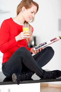 Young woman sat on kitchen work top, enjoying a glass og juice and reading a magazine