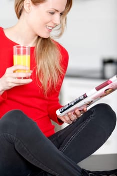 Young woman sat on kitchen work top, enjoying a glass og juice and reading a magazine