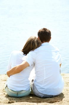 Backs of serene couple sitting on sandy shore in front of blue water