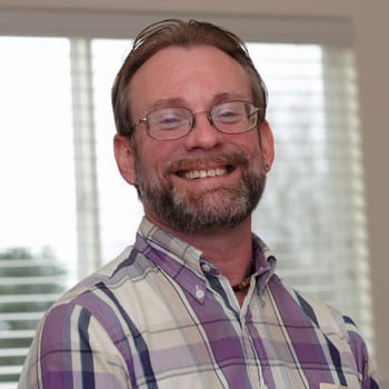 Male wearing eyeglasses in is forties with blonde gray hair and beard smiling in a joyous way showing his upper teeth.