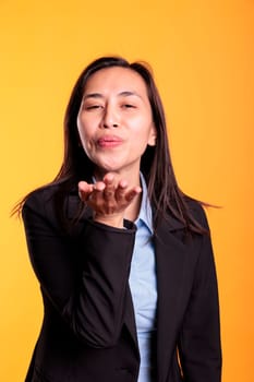 Attractive asian model blowing air kissed during studio shot, posing over yellow background. Confident carefree woman expressing love, looking at the camera with a satisfied smile. Romantic gesture