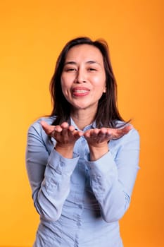 Cheerful brunette filipino woman blowing air kissed during studio shot, posing over yellow background. Positive young adult expressing love, looking at the camera with a flirty smile. Romantic gesture