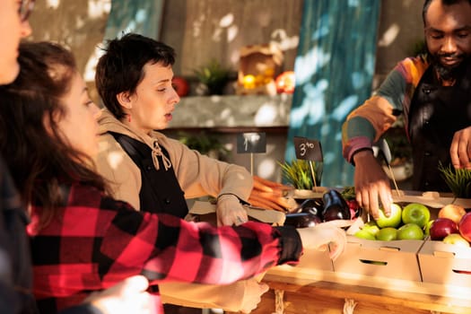 Young vendor helping client to choose fruits and vegetables, selling fresh organic homegrown produce at local farmers market. Woman talking to seller and buying natural seasonal food.